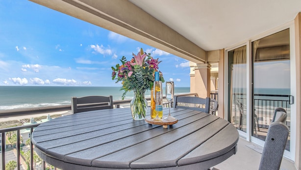 Une table ronde ornée d’un vase de fleurs et de deux bouteilles, placée sur un balcon donnant sur l’océan.