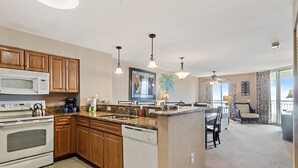 Una cocina con armarios de madera, microondas, estufa y lavavajillas. Hay una zona de comedor con mesa y sillas, y vistas al exterior a través de grandes ventanales.