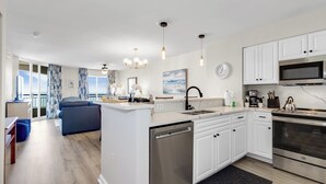 A modern kitchen with white cabinets, a dishwasher, a microwave, and an oven. There is a dining area with a blue sofa and a chandelier.