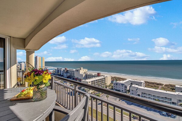 Un balcon avec vue sur la plage et l’océan, une table ornée d’un plateau de fruits et de fleurs, et un paysage urbain en arrière-plan.