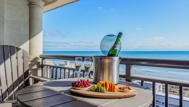 A balcony with a table set for a meal, a bottle of champagne, and a view of the ocean. - Newly Refreshed & Cozy N. Myrtle Beach Condo with 15th Floor Ocean views (North Myrtle Beach)