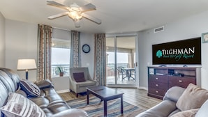 Een moderne woonkamer met een leren bank, een salontafel en een televisie op een standaard. Er is een plafondventilator, een raam met gordijnen en uitzicht op het balkon.