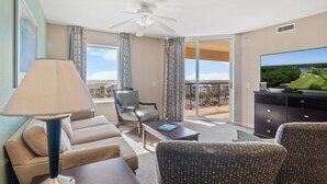 Ein Hotelzimmer mit einer Couch, einem Sessel und einem Fernseher auf einer Kommode. Ein Balkon bietet Blick auf einen Golfplatz und Bäume.