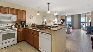 A modern kitchen with granite countertops, wooden cabinets, a built-in oven, and a dishwasher. There is a dining area with a view of the outdoors.