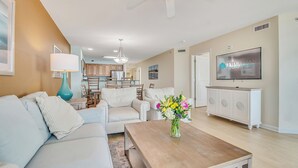 Een woonkamer met een bank, salontafel en televisie. Er is een plafondventilator, een lamp en een vaas met bloemen.