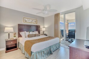 Une chambre d’hôtel avec un grand lit, deux tables de chevet avec des lampes, un ventilateur de plafond, un bureau avec une chaise et une vue sur l’océan par une porte-fenêtre coulissante.