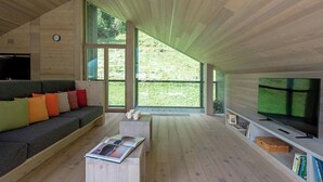 Een moderne woonkamer met een houten plafond, grote ramen, een bank met kleurrijke kussens, een salontafel met tijdschriften en een televisie op een dressoir.