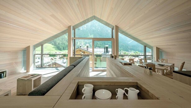 A modern interior with a sloped ceiling, large windows offering a view of a mountainous landscape, a dining area with a wooden table and chairs, and a built-in seating area with a bench and a coffee table.