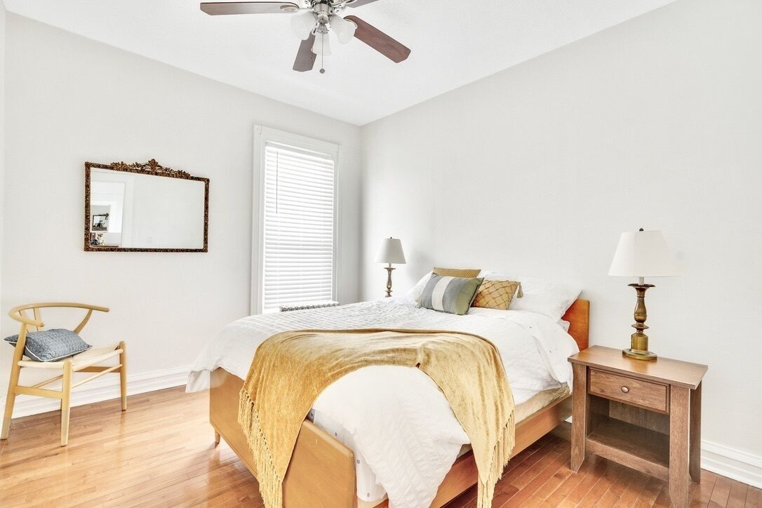A bedroom with a bed, bedside tables, a ceiling fan, a window with blinds, a chair, and a mirror.