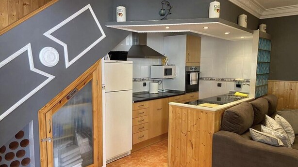 Eine kompakte Küche mit eingebautem Herd, Mikrowelle und Backofen. Es gibt einen Holzausbau mit Glastür und eine kleine Kücheninsel mit Spüle. Die Küche hat ein modernes Design mit weißen und grauen Fliesen.
