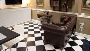 Ein Wohnzimmer mit schwarz-weiß kariertem Boden, einer braunen Ledersofa, einem Fernseher, der 'NETFLIX' zeigt, und einem Bücherregal mit verschiedenen Gegenständen.