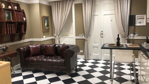 Ein Zimmer mit Schachbrettmuster-Boden, einem Ledersofa, einem Esstisch mit Stühlen, einem Bücherregal und einer Tür mit Vorhängen.