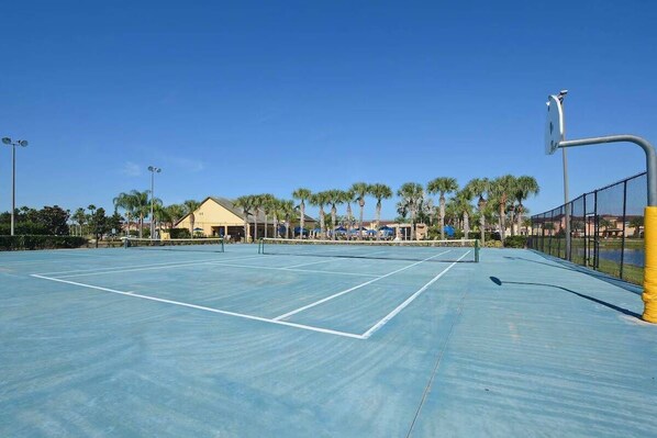 An outdoor tennis court with a net, surrounded by a fence and palm trees.