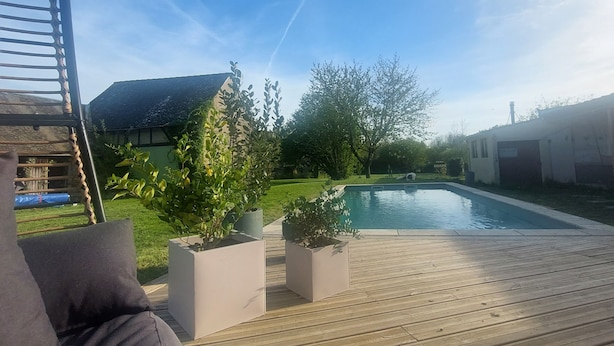 Une terrasse en bois avec une piscine, des plantes en pot et une vue sur une maison dont le toit est en pente.