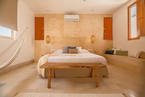 Une chambre dotée d’un grand lit, d’un banc en bois, de deux luminaires suspendus, d’un appareil de climatisation et d’une fenêtre offrant une vue agréable.