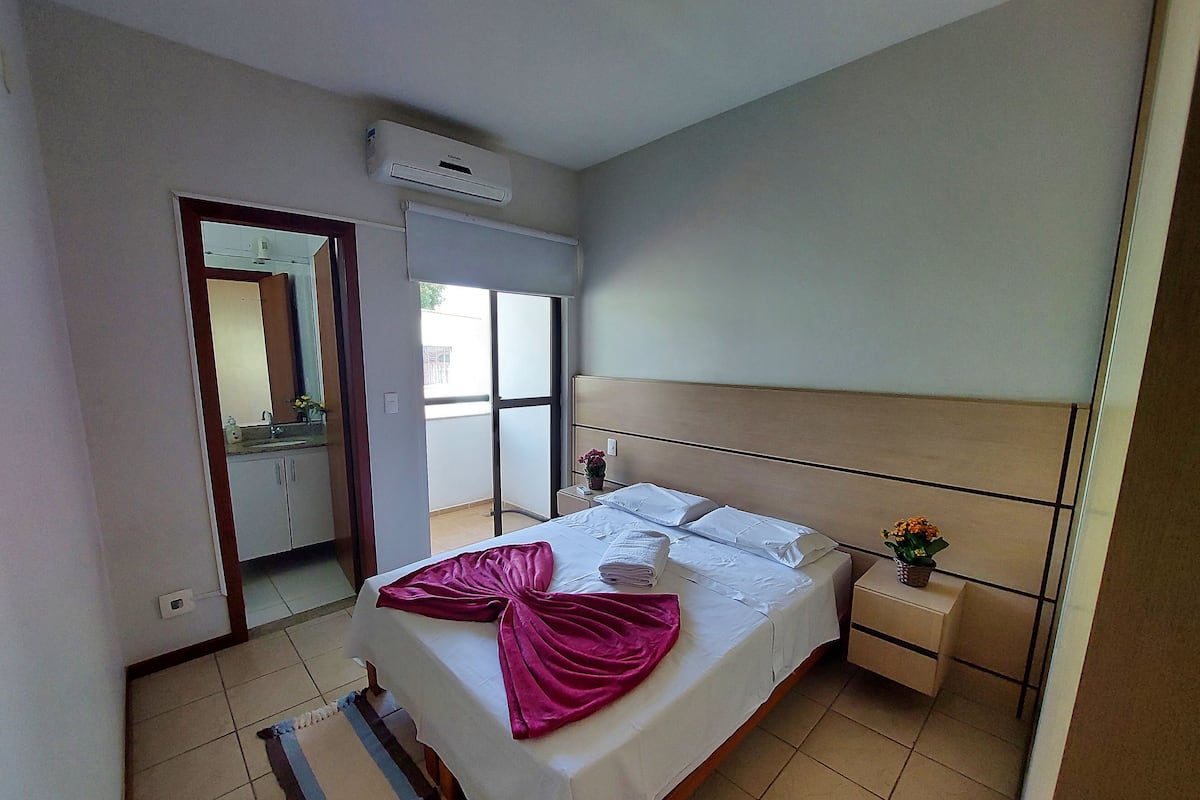 A hotel room with a bed, a nightstand, a mirror, an air conditioning unit, and a view of the outside.