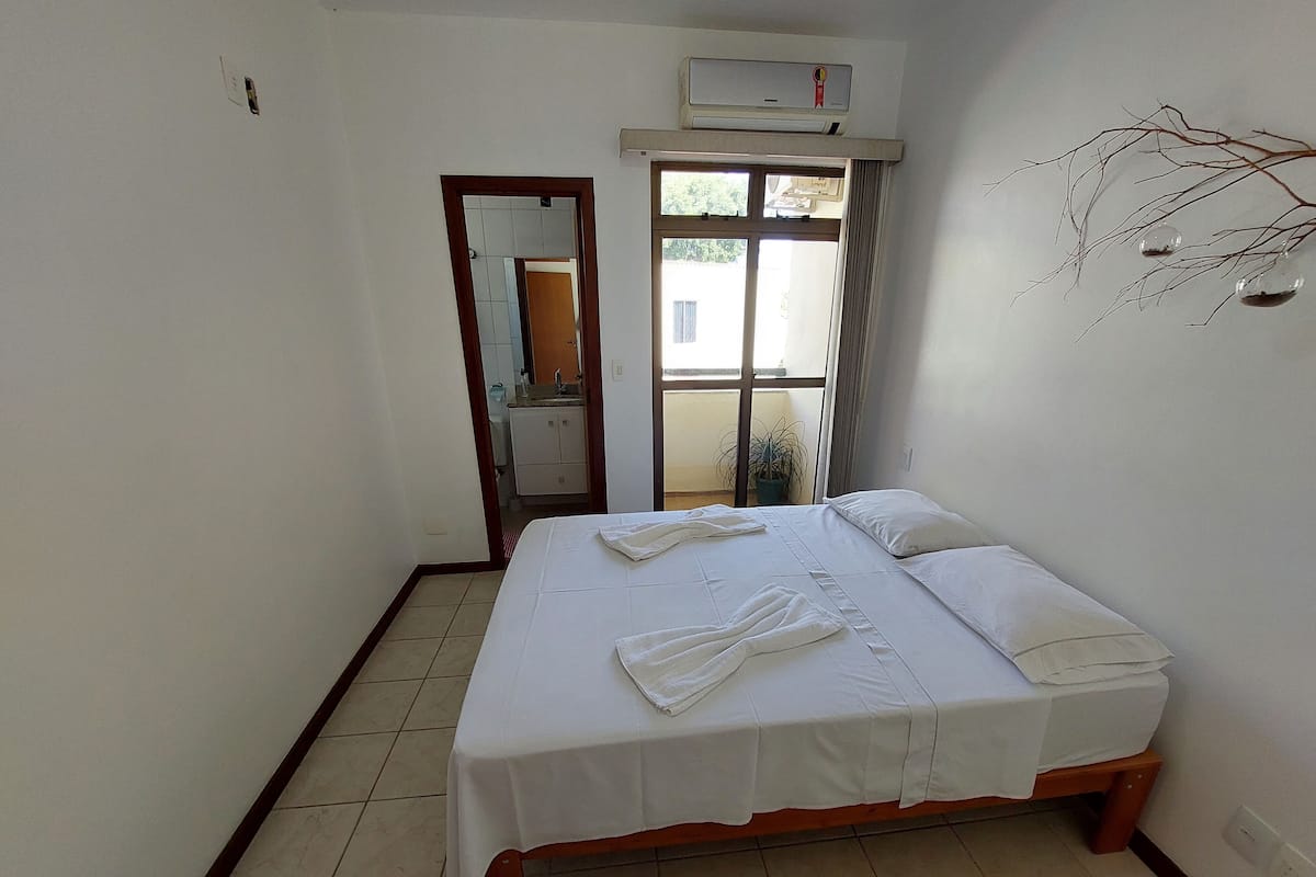 A neatly made bed with white linens and towels, a wooden headboard, a window with curtains, and a wall-mounted air conditioner.