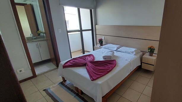 A hotel room with a bed, a nightstand, a small table with a vase of flowers, and a sliding door leading to a balcony. - Condomínio Santa Mônica (Belo Horizonte)