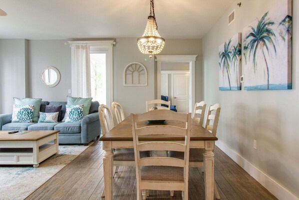 A dining area with a wooden table and chairs, a chandelier, and a painting of palm trees on the wall.