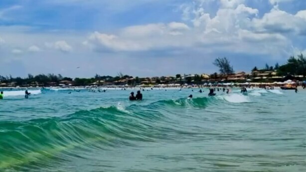 A beach with clear skies and waves, with people surfing and swimming. - Pousada Cielo Blu (Búzios)