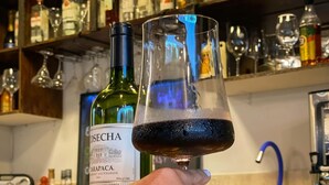 A hand holding a wine glass with a dark liquid, a bottle of Josecha wine, and a bar with shelves of various bottles and glasses. - Pousada Cielo Blu (Búzios)