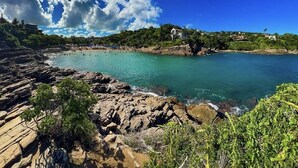 A coastal landscape with rocky shores, clear turquoise water, and buildings on a hillside. - Pousada Cielo Blu (Búzios)