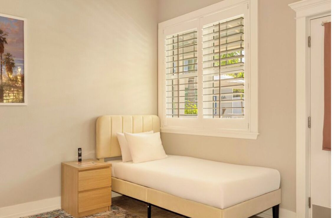A bedroom with a bed, a nightstand, a window with blinds, and a painting on the wall.