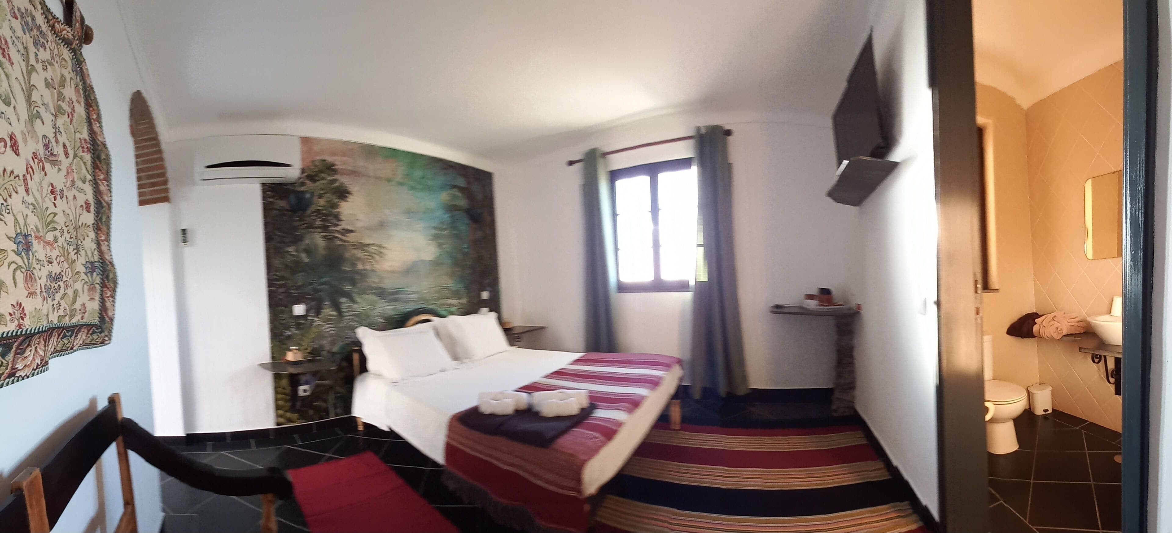 Una camera d'albergo compatta con un letto, una piccola scrivania, una finestra con tende, un bagno con il water e il lavandino e un arazzo a parete.