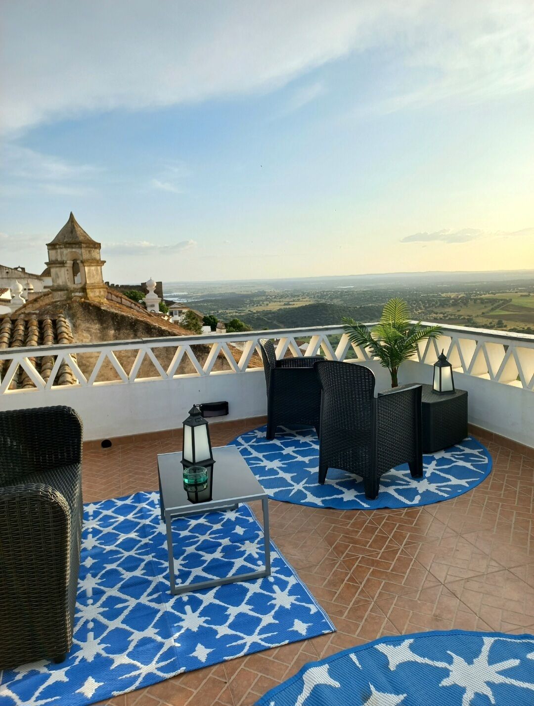 A rooftop terrace with wicker furniture, a table, and a view of a distant cityscape and fortifications.