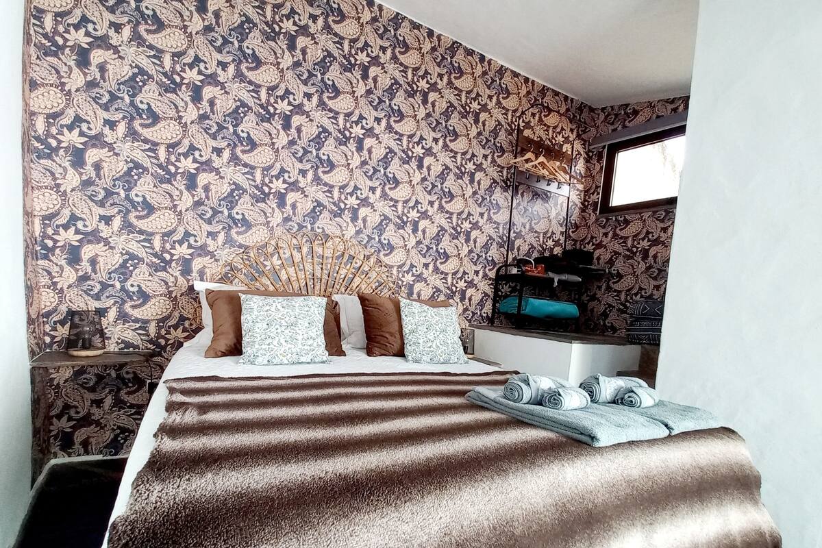 Un dormitorio con papel tapiz estampado, una cama con manta a rayas, almohadas, una mesita de noche con lámpara y una ventana con vistas.