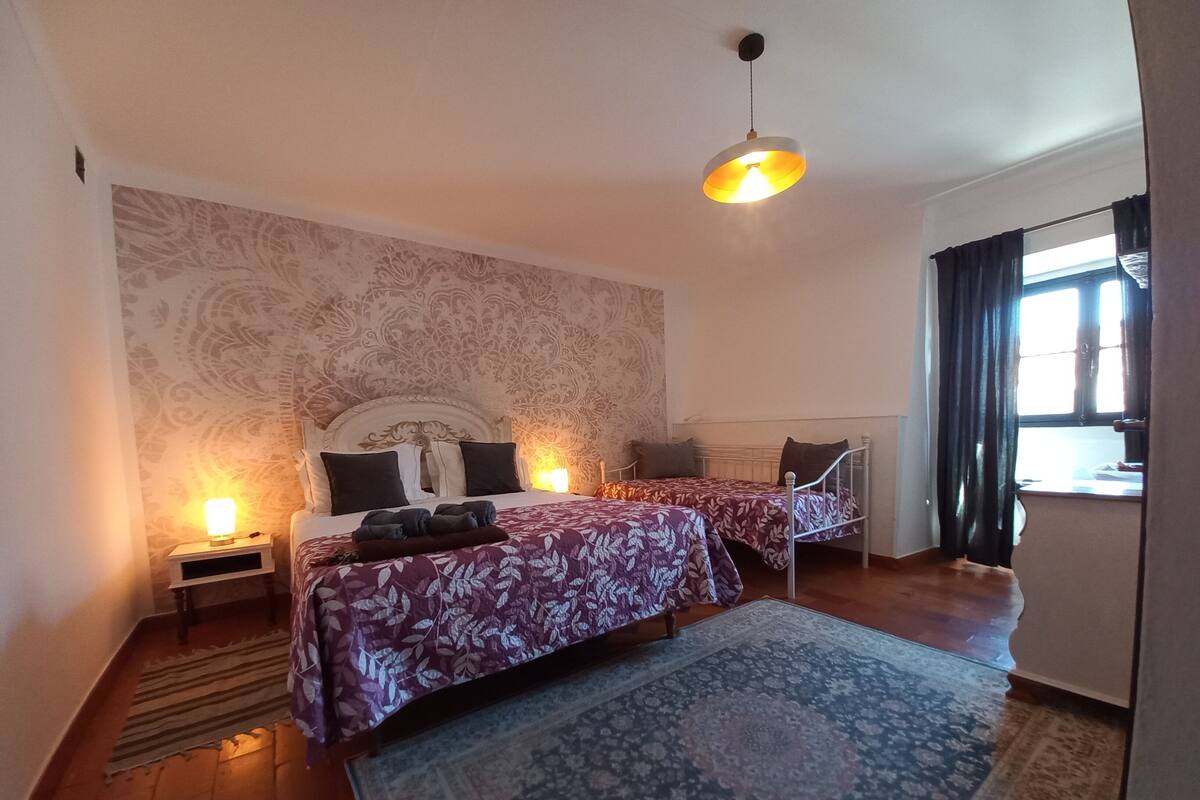 Un dormitorio con dos camas, un papel tapiz decorado, una lámpara colgante y una ventana con cortinas.