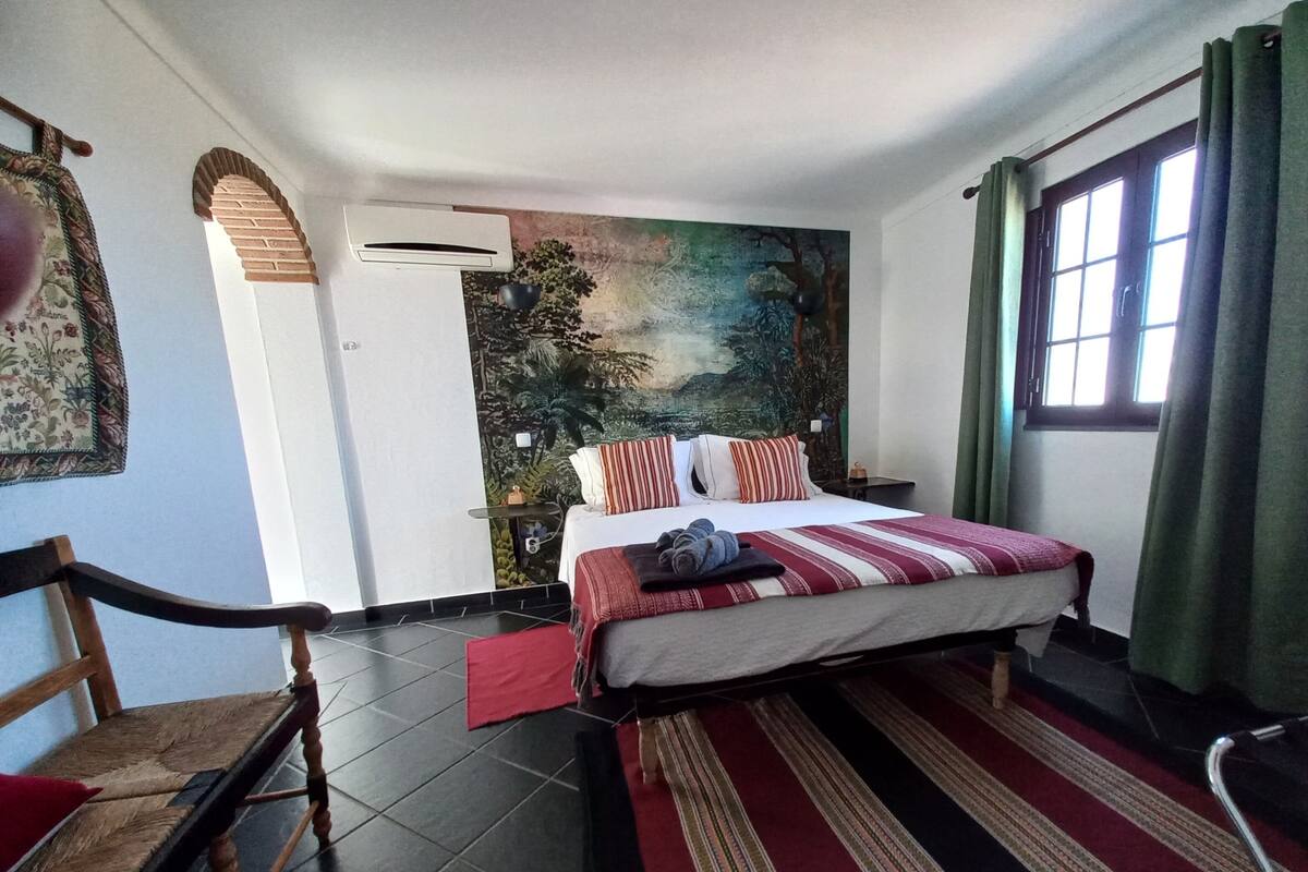 Un dormitorio con cama, una silla, una ventana con cortinas, un aire acondicionado instalado en la pared y un mural.