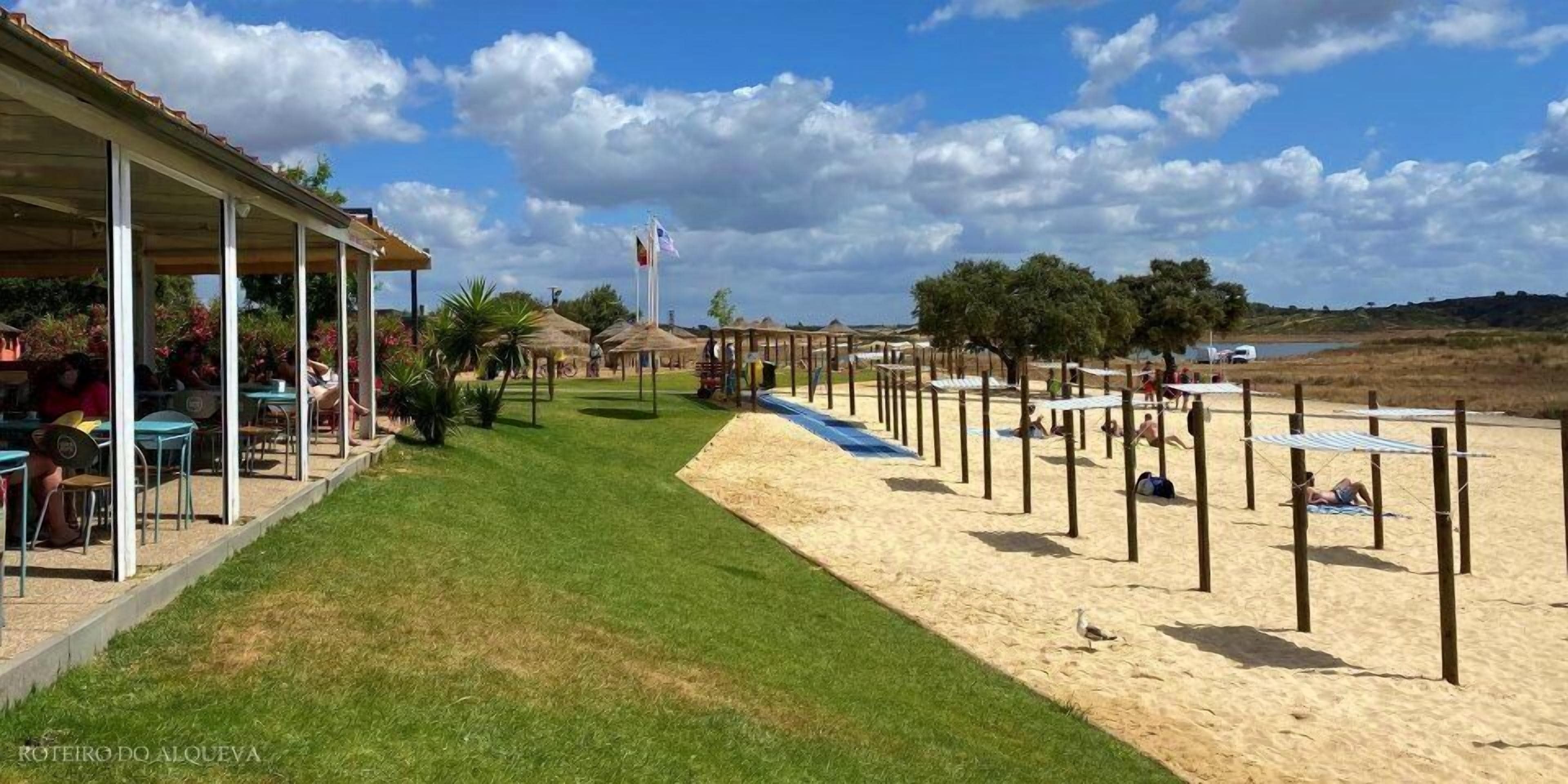 A beachside area with a covered seating area, a sandy beach with umbrellas, and a grassy path.