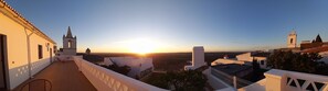 A terrace with a view of a sunset over a valley, featuring white buildings with distinct architectural styles. - Casa Dona Antonia - Monsaraz (Reguengos de Monsaraz)