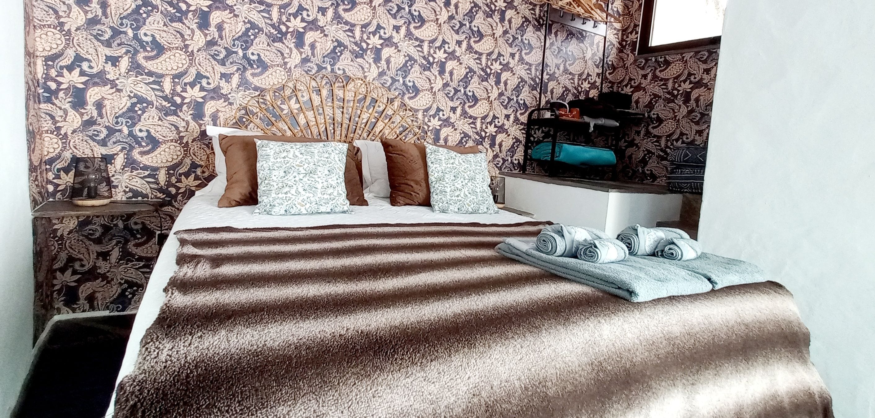 Un dormitorio con papel tapiz estampado, una cama con manta a rayas, almohadas y una mesita de noche con una lámpara y un objeto azul.