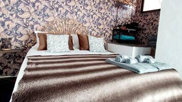 Un dormitorio con papel tapiz estampado, una cama con manta a rayas, almohadas y una mesita de noche con una lámpara y un objeto azul.