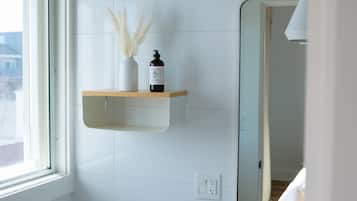 A bathroom with a white tiled wall, a wooden shelf with a bottle and a vase, and a mirror.