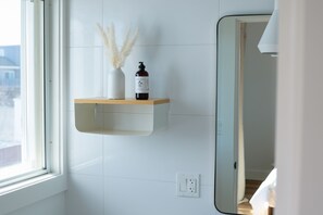 A bathroom with a white tiled wall, a wooden shelf with a bottle and a vase, and a mirror. - DUNE Fairfield Beach (Fairfield)