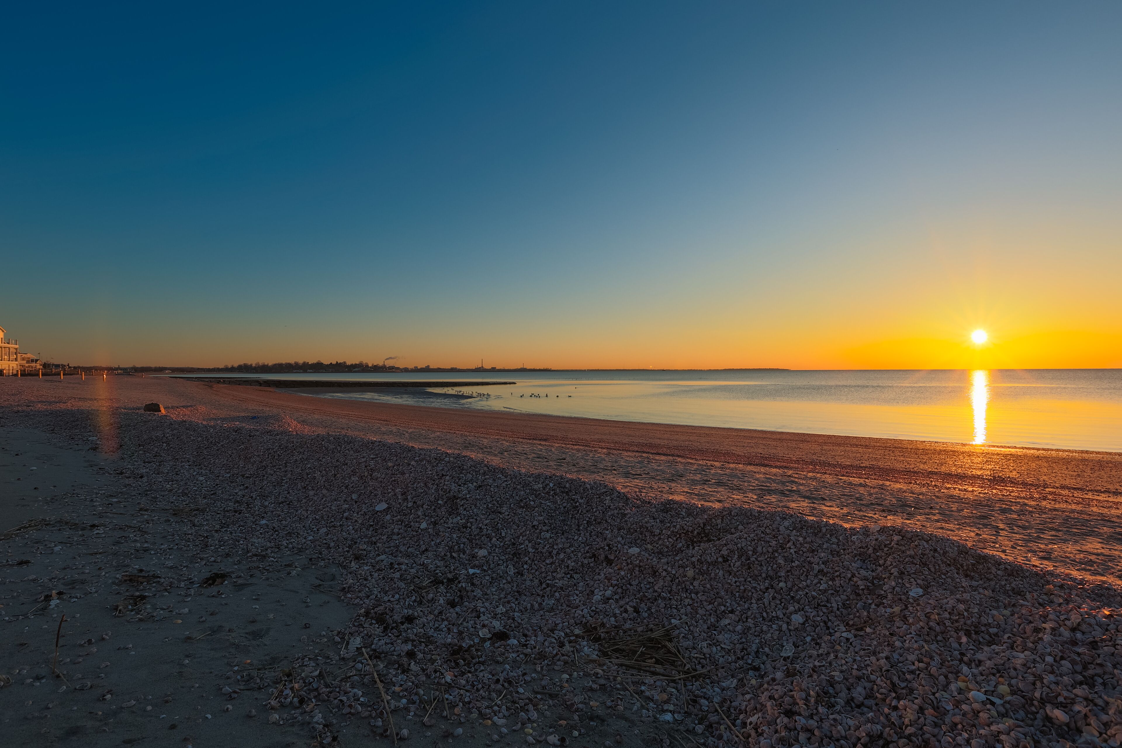 A pebbled beach at sunset with a calm sea and a clear sky.