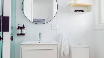 Um banheiro moderno com pia branca, espelho e vaso sanitário. Há uma toalha pendurada na parede e um cesto de lixo pequeno no chão.