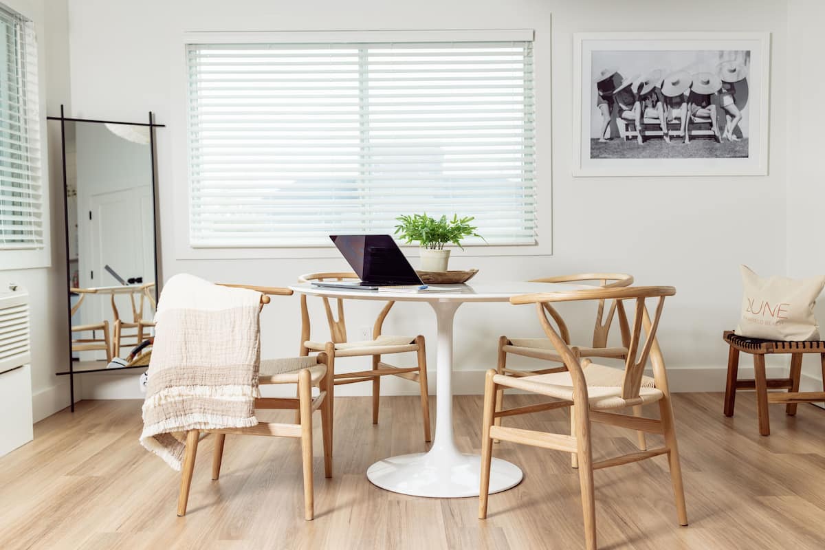 Une pièce moderne avec une table ronde blanche, quatre chaises en bois, un ordinateur portable, une plante en pot et une photographie en noir et blanc encadrée accrochée au mur.