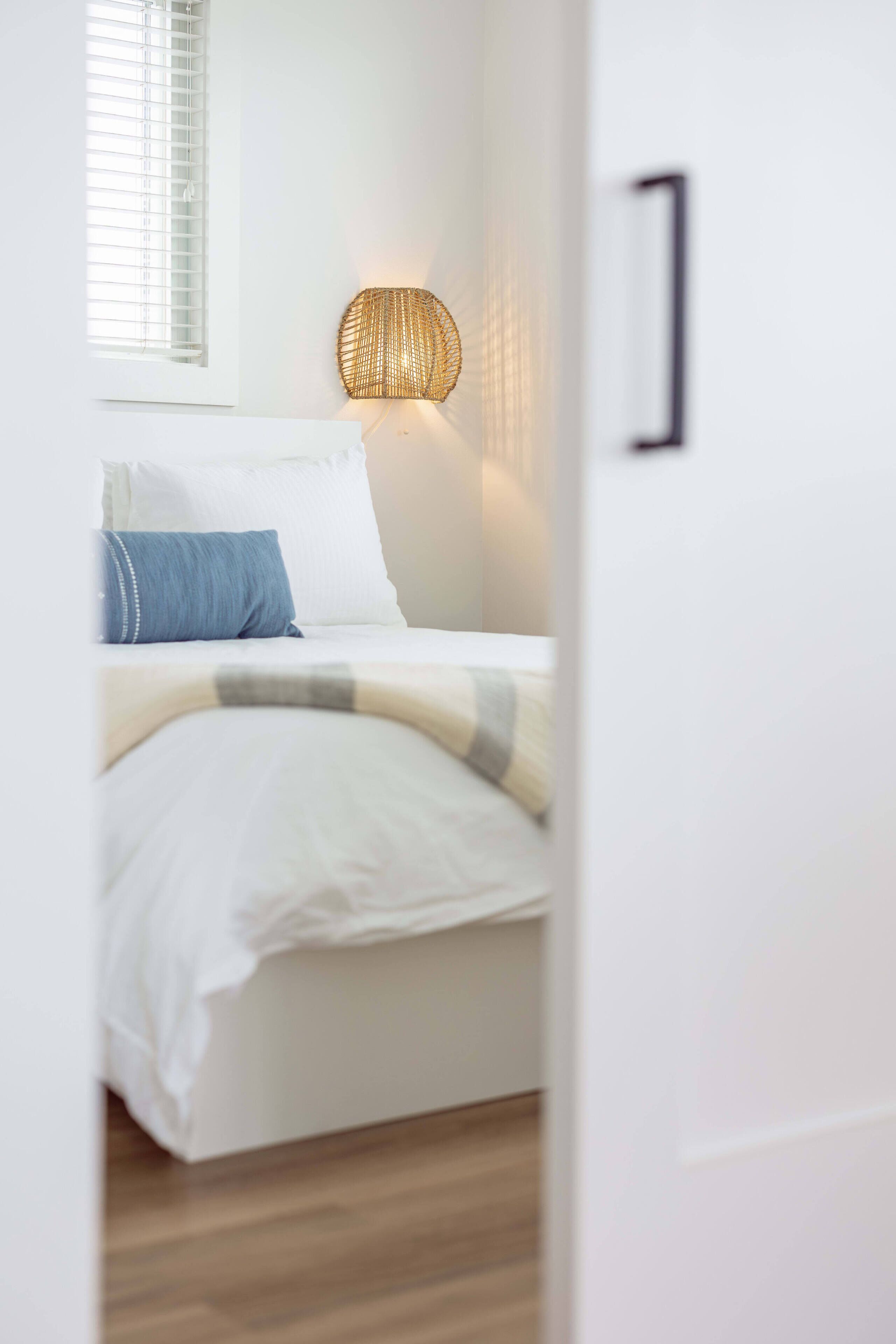 Une chambre à coucher avec un lit, du linge de lit blanc, un oreiller bleu, une lampe murale en osier et une fenêtre avec des stores.