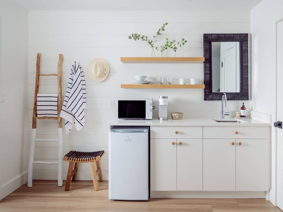 A compact kitchen with white cabinetry, a microwave, a small refrigerator, and wooden shelves above the counter.
