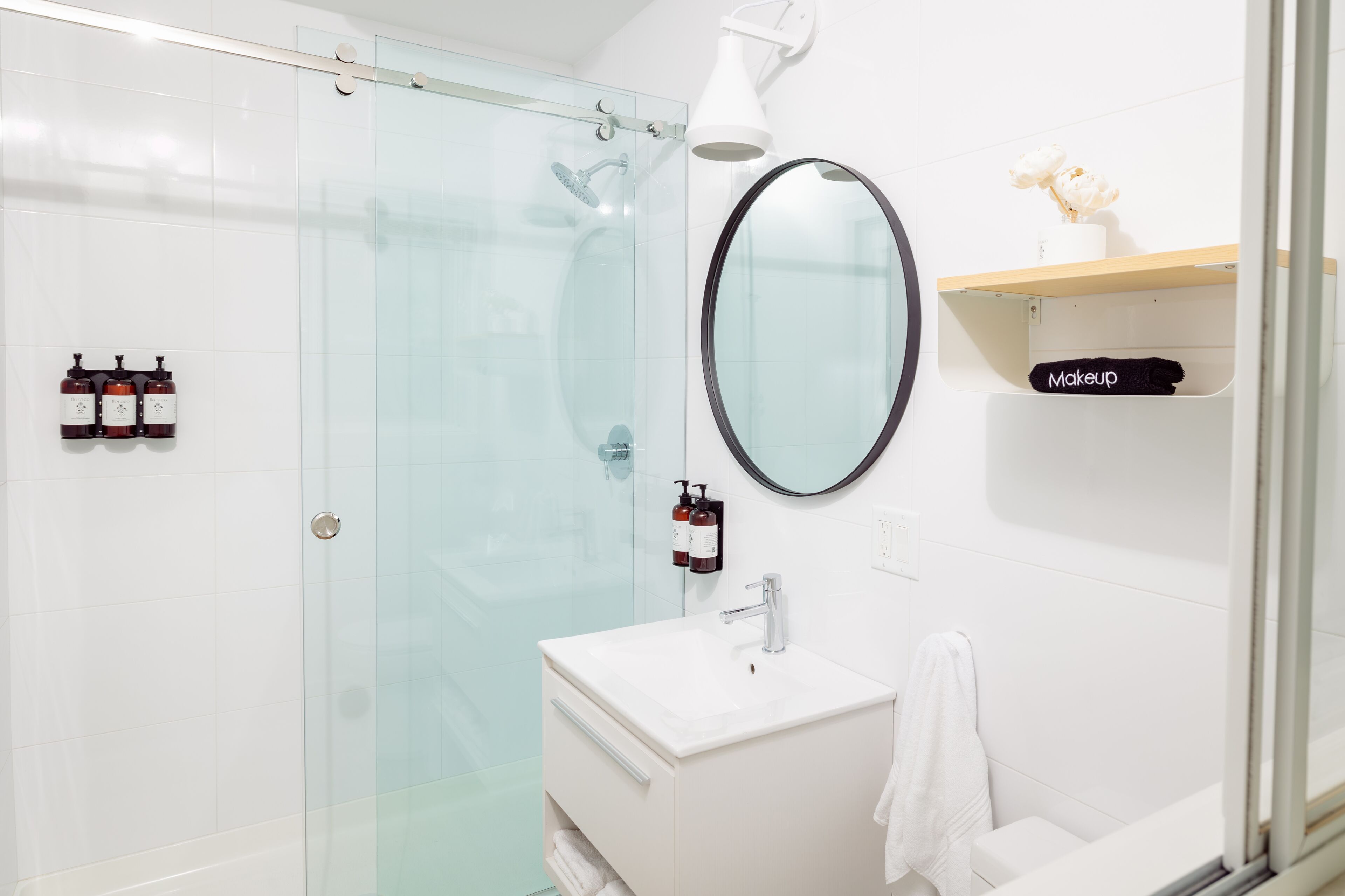 Un baño moderno con cabina de ducha de vidrio, un lavamanos blanco, un espejo y una repisa con un cosmético.