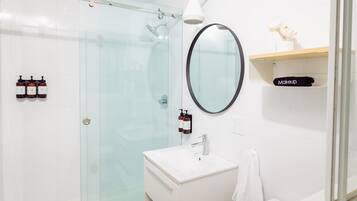 Un baño moderno con cabina de ducha de vidrio, un lavamanos blanco, un espejo y una repisa con un cosmético.