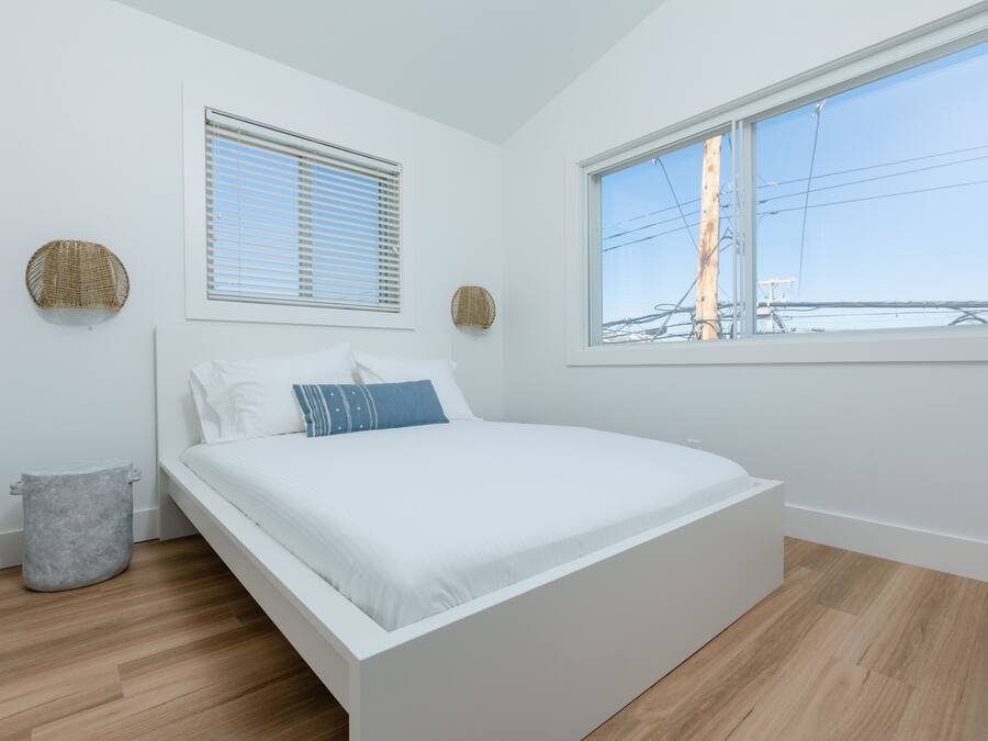 A bedroom with a white bed, two windows, and a grey trash bin.