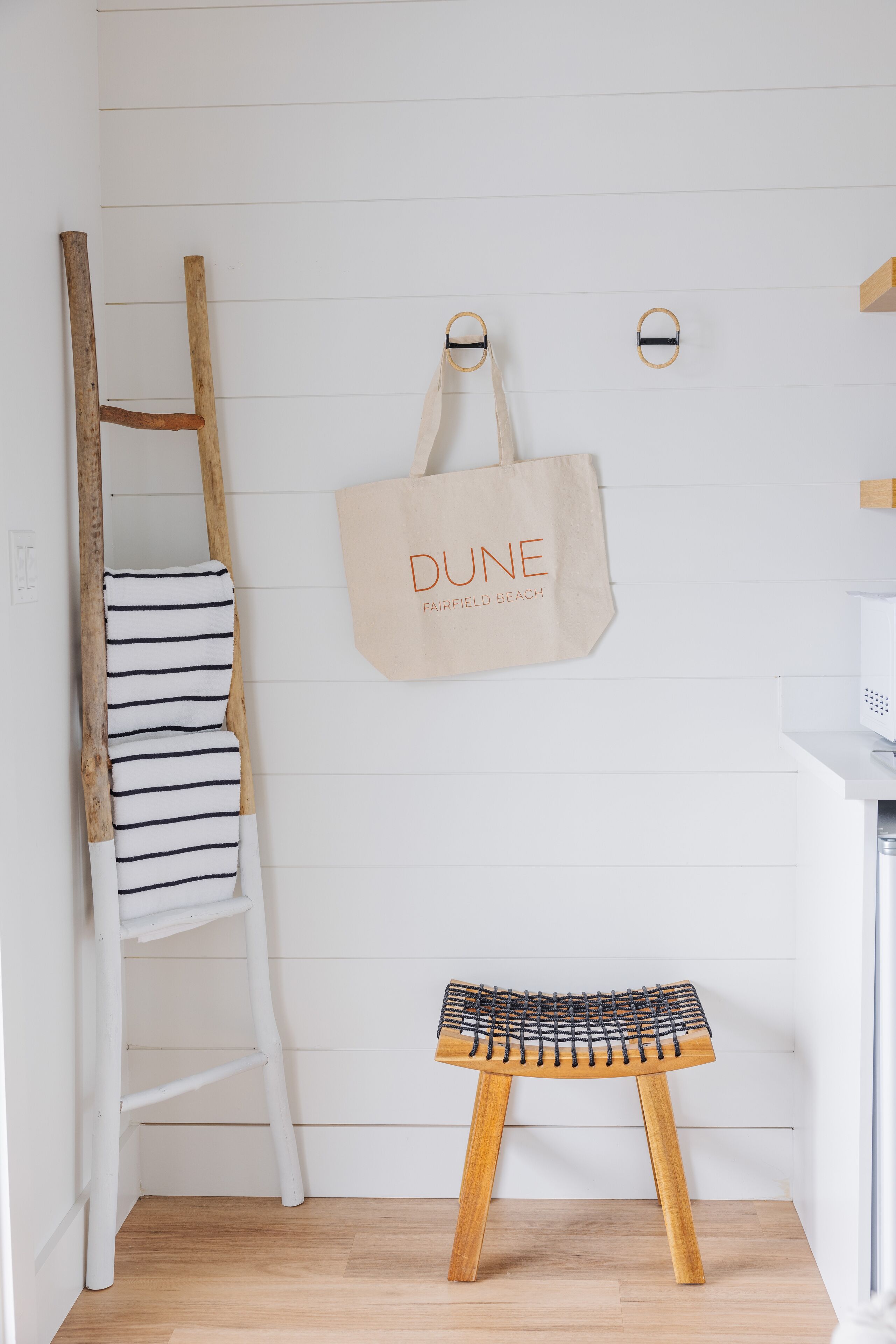 A wooden ladder leaning against a white wall with a striped cushion and a small stool. A beige tote bag hangs on the wall.