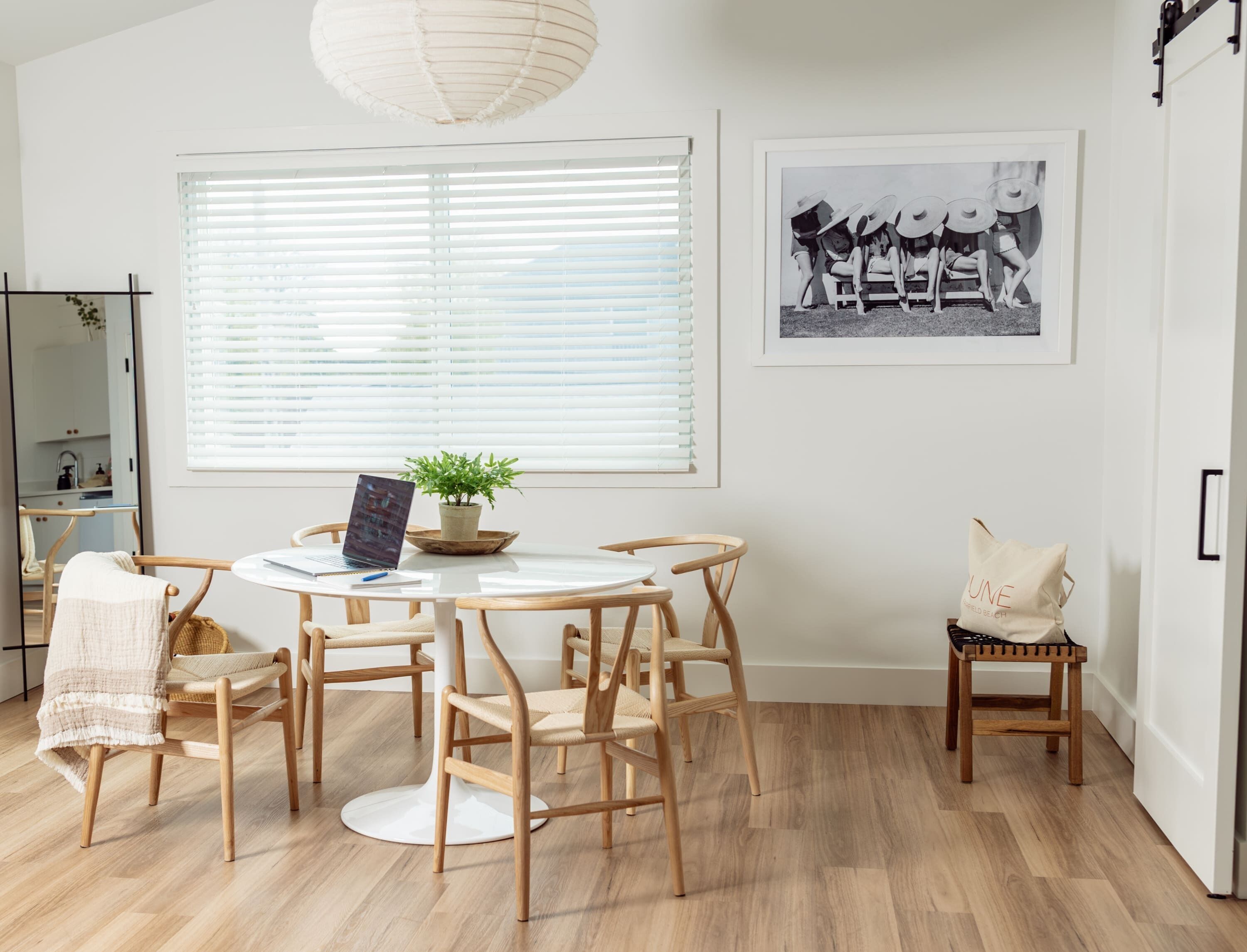 A dining area with a round table, wooden chairs, a potted plant, and a large window with blinds.