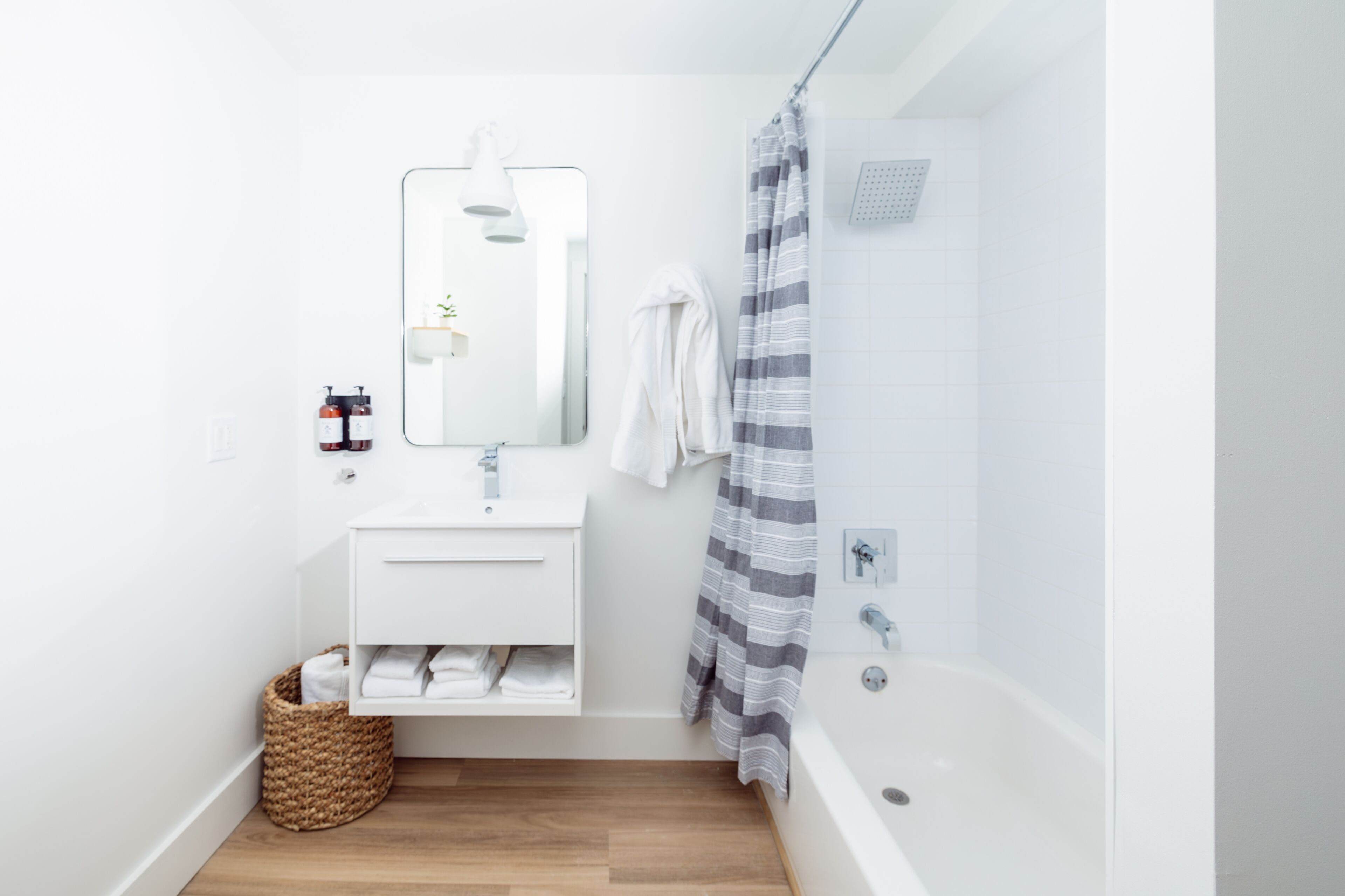 Un baño moderno con un tocador blanco, un espejo grande, una ducha con cortina a rayas y suelo de madera.
