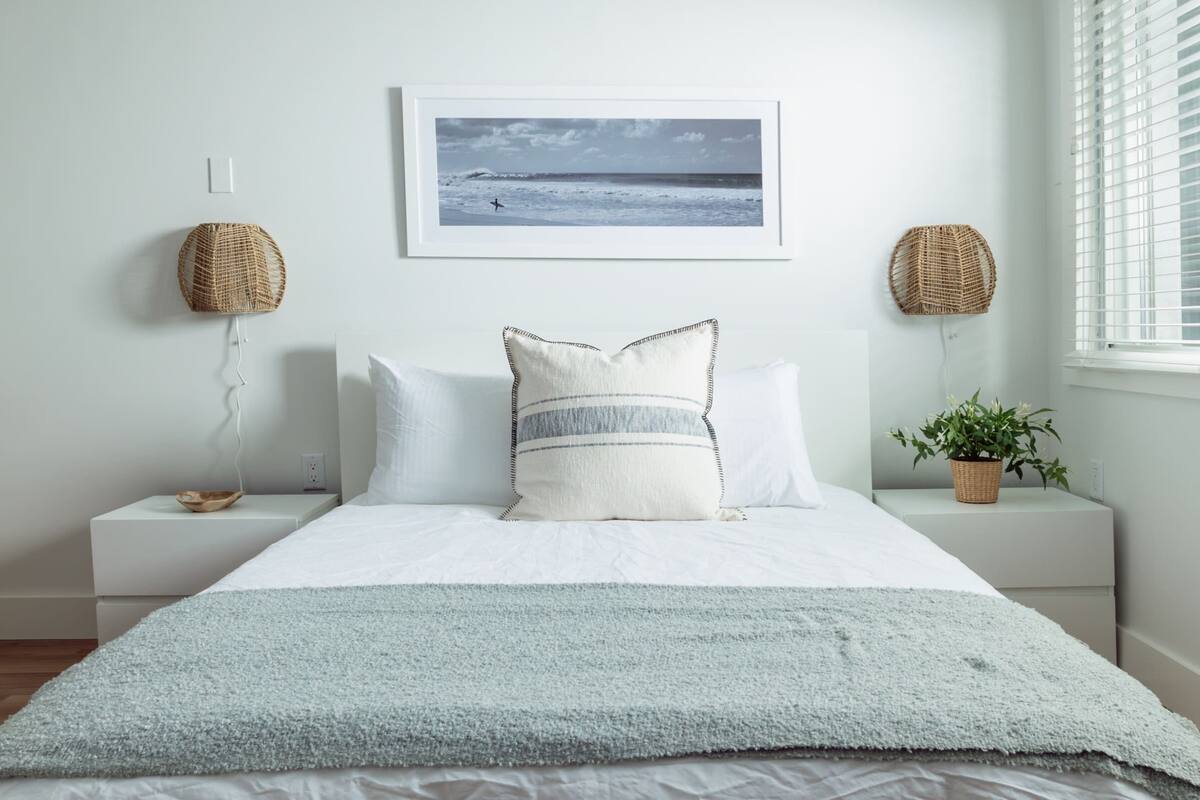 Une chambre à coucher avec un lit, du linge de lit blanc, un tableau encadré au mur, deux tables de chevet avec des lampes en osier, une fenêtre avec des stores et une petite plante en pot.